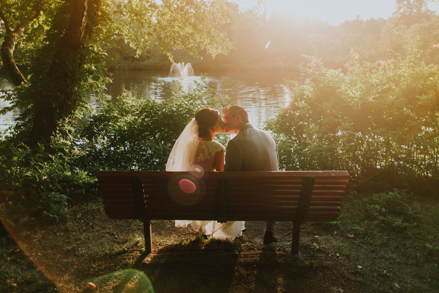 Kissing Bride and Groom on a Park Bench by a Lake and Fountain with Shining Sunlight | Confetti.co.uk